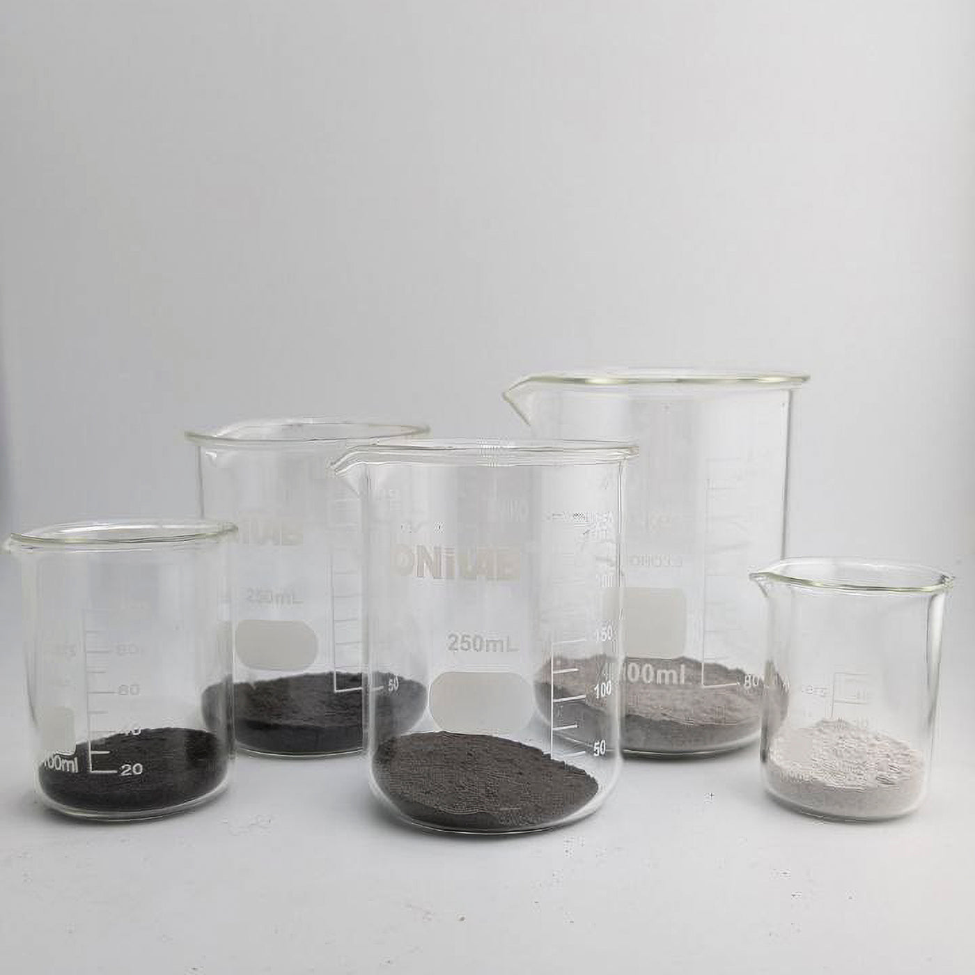 Set of clear glass beakers with different pigments ranging from black to white on a white background.