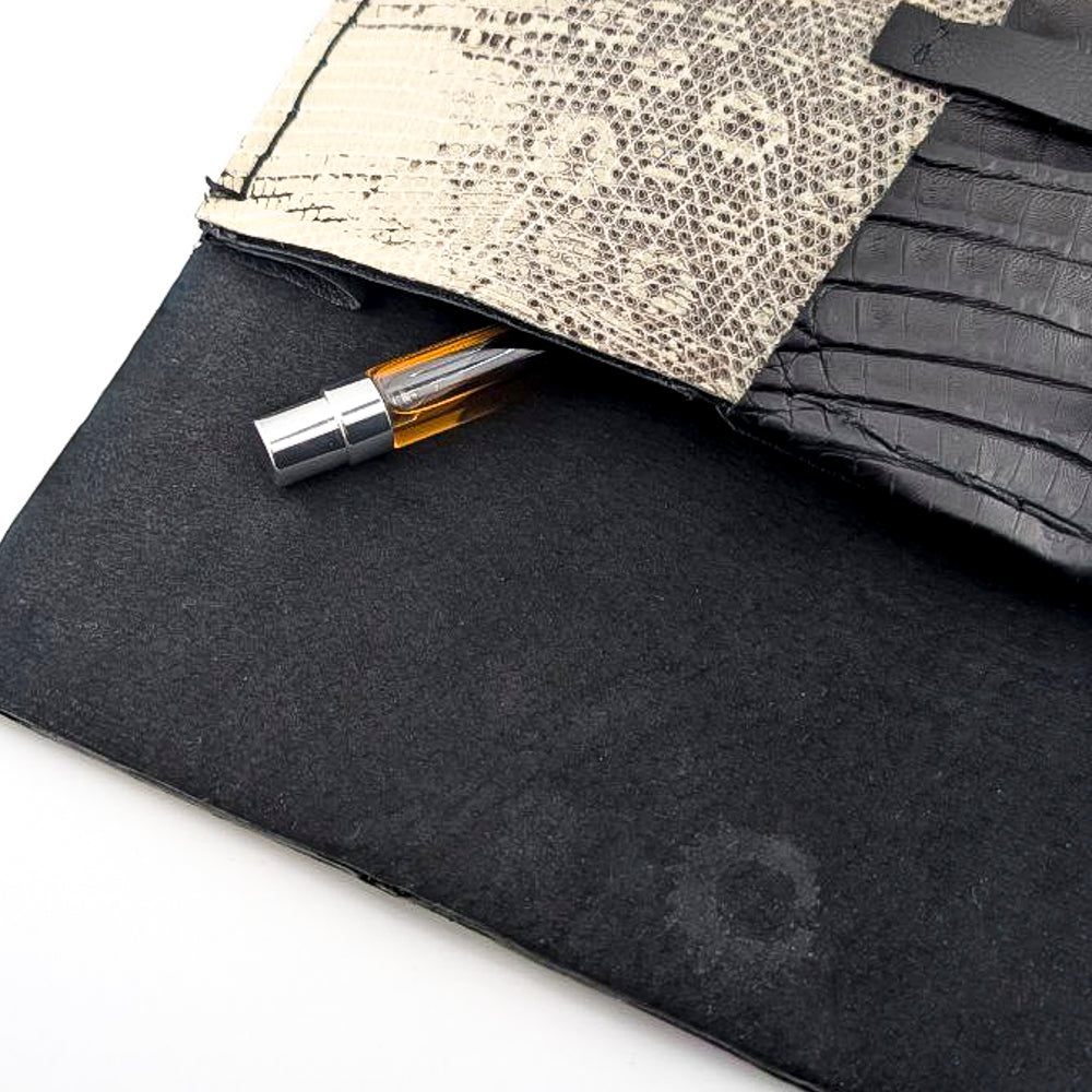 Black and white lizard leather clutch with amber perfume vial against a white background