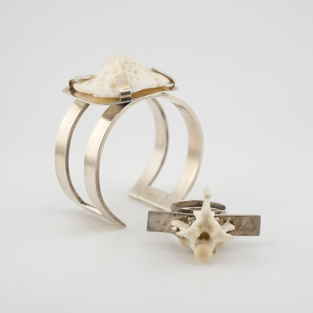 A large silver arm cuff with bone set into t paired next to a chunky silver ring with vertebrae against a white background.