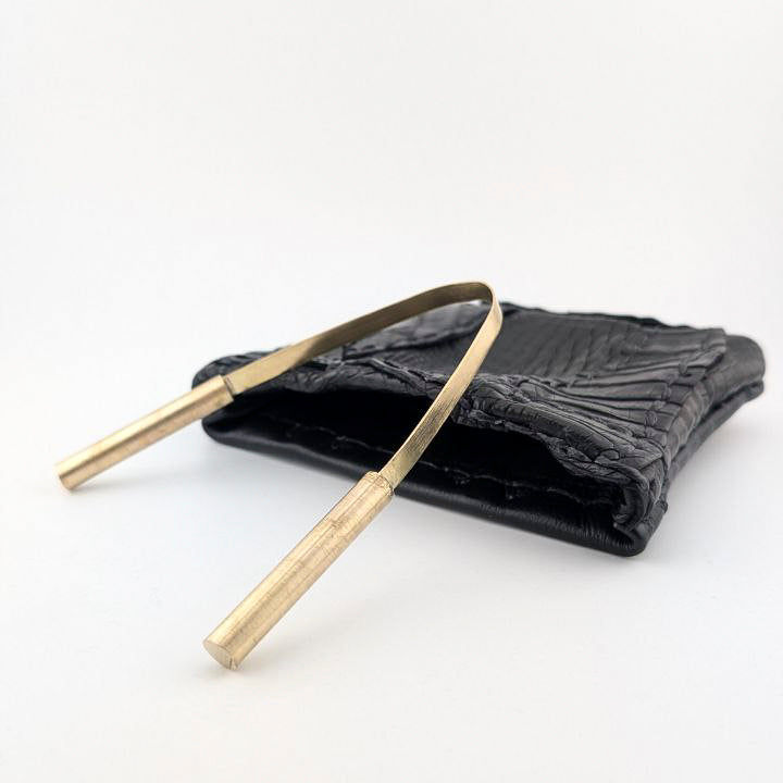 Black textured pouch with brass tongue scraper on a white background